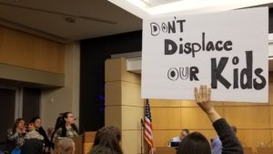 Virginia Herrera-Páramo addresses the council while a woman raises a sign that reads Don't displace our kids