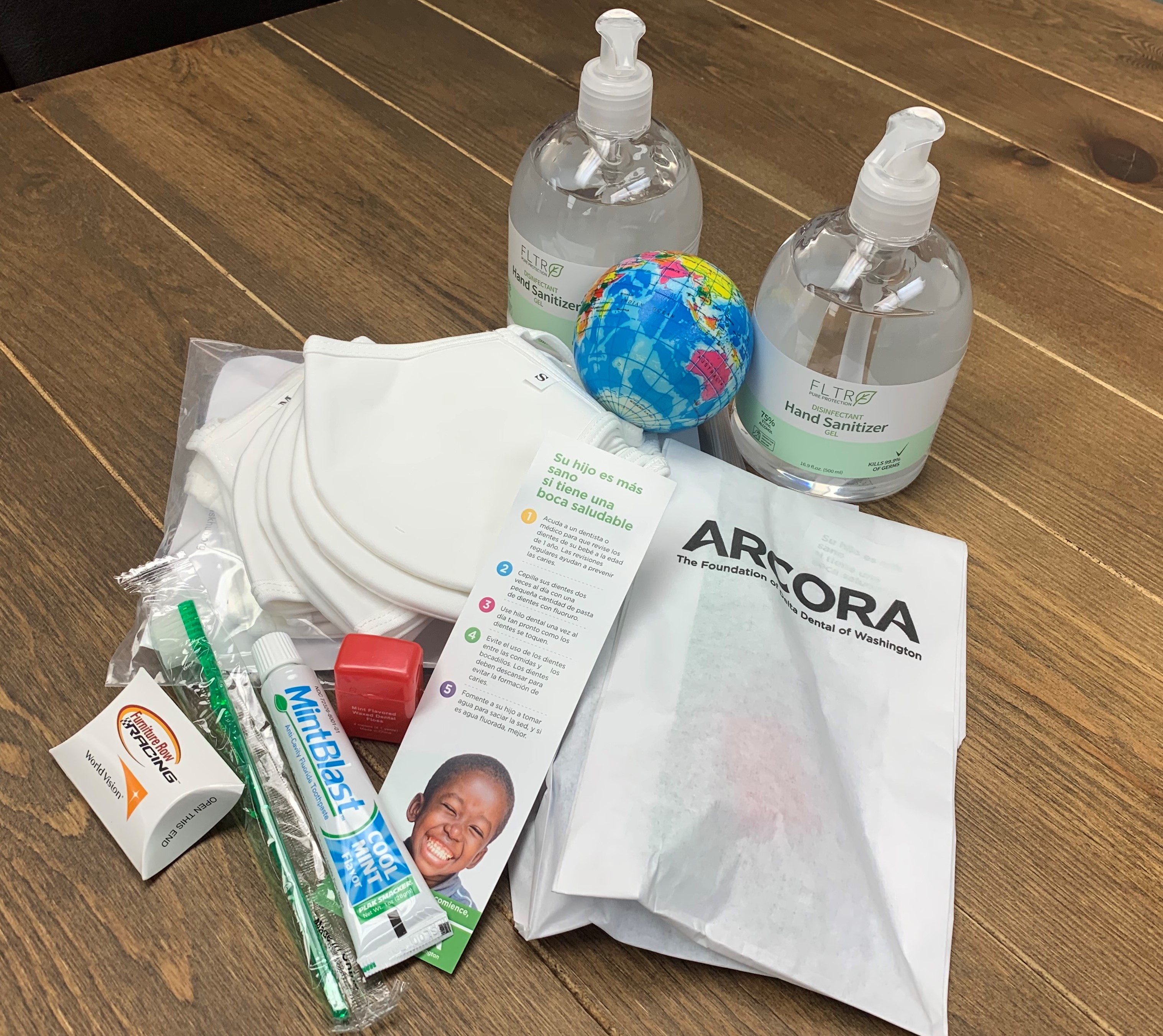 On a wooden table there is hand sanitizers, a stress ball, face masks, and a dental kit from Arcora Foundation
