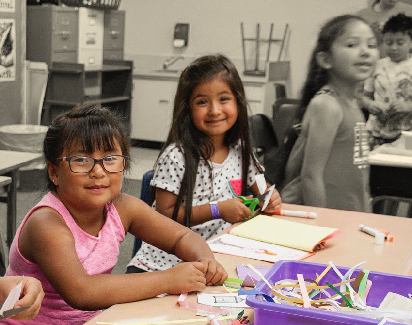 Two young girls sit at a table doing crafts smiling at the camera while their background and other children are in black and white and blurred.
